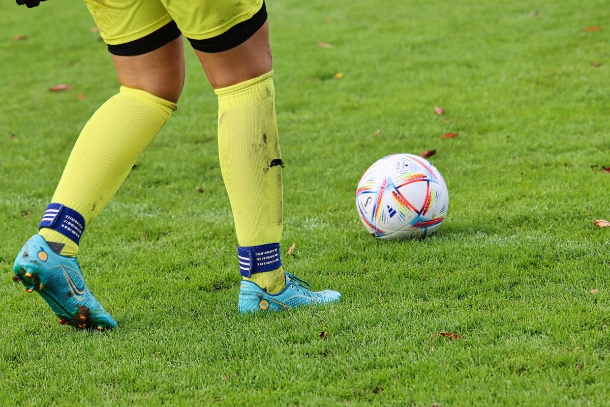 Ein Fußballspieler in gelben Socken steht auf einem Rasenfeld mit einem bunten Fußball in der Nähe.
