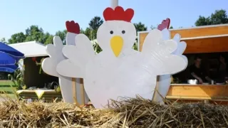 Eine große weiße Holzskulptur einer Henne mit roten Kämmen und gelbem Schnabel, die auf einem Heuhaufen sitzt.