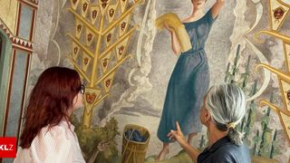 Zwei Frauen betrachten ein Wandbild, das eine Frau mit einem Korb Trauben zeigt. Das Wandbild zeigt einen goldenen Baum mit dreieckigen Emblemen und einem bewölkten Himmel.