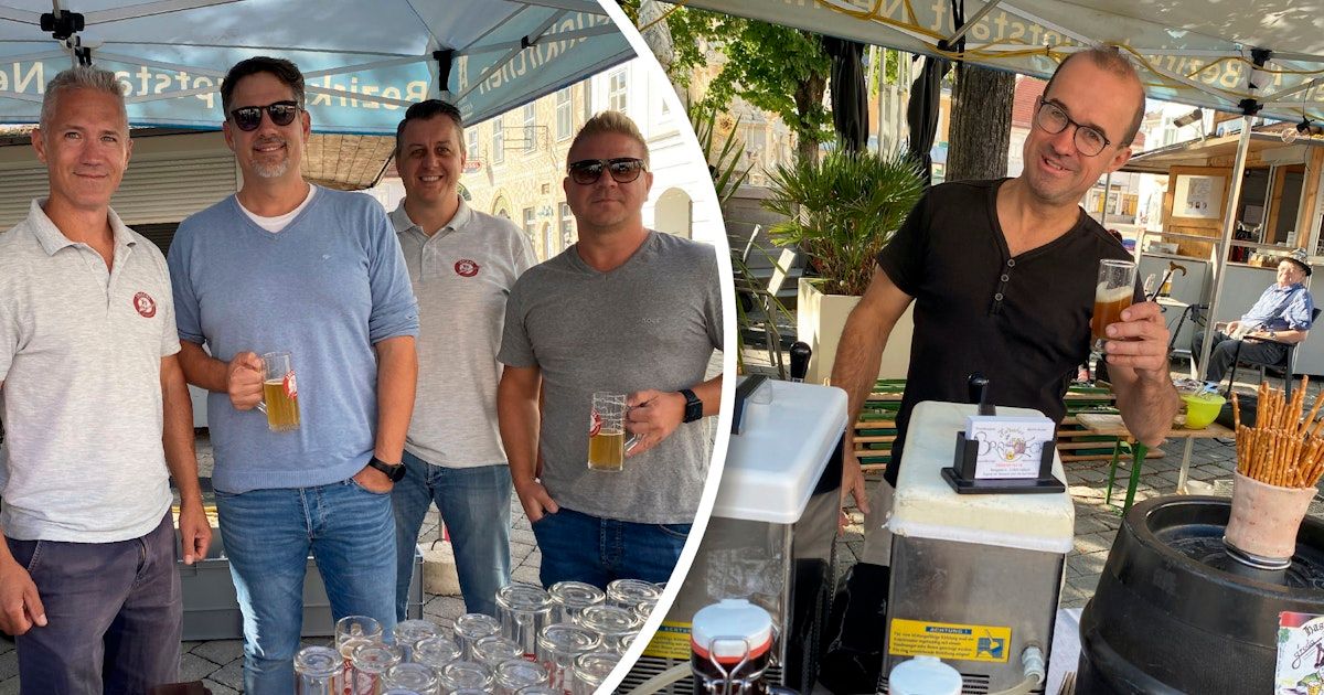 Vier Männer stehen unter einem Schirm, lächeln und halten Gläser mit Bier in der Hand. Zwei von ihnen tragen Sonnenbrillen. Im Hintergrund ist ein verschwommenes Bild einer Bar mit einem Bierhahn und einem Topf zu sehen.
