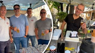 Vier Männer stehen unter einem Schirm, lächeln und halten Gläser mit Bier in der Hand. Zwei von ihnen tragen Sonnenbrillen. Im Hintergrund ist ein verschwommenes Bild einer Bar mit einem Bierhahn und einem Topf zu sehen.