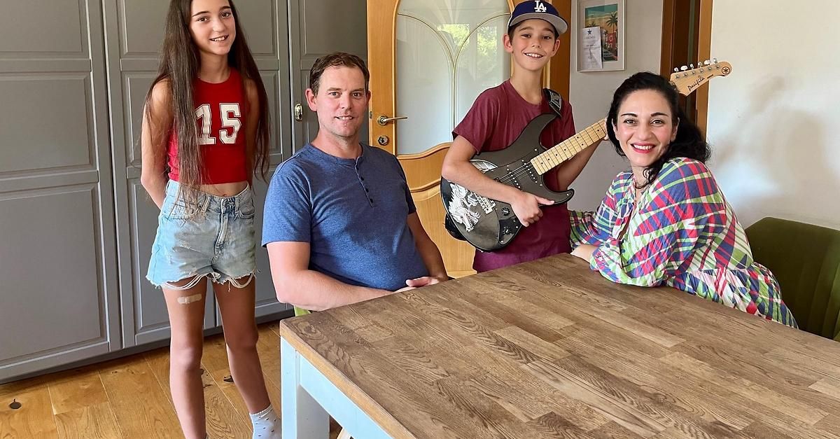 Eine vierköpfige Familie posiert für ein Foto in einem Raum mit einem Holztisch. Der Mann sitzt und die Frau steht neben ihm. Der Junge hält eine Gitarre und das Mädchen steht neben ihm.