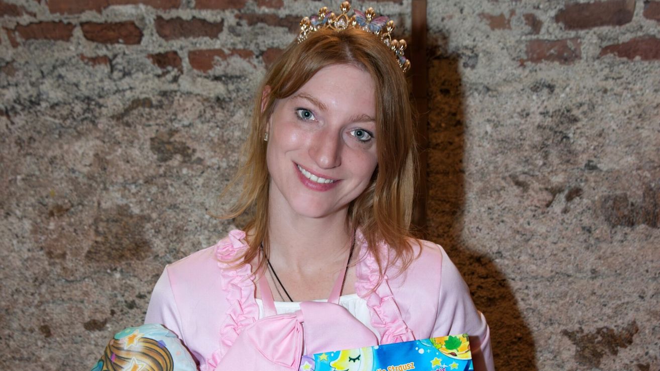 Eine Frau in einem rosa Kleid und Krone lächelt und hält ein Buch.