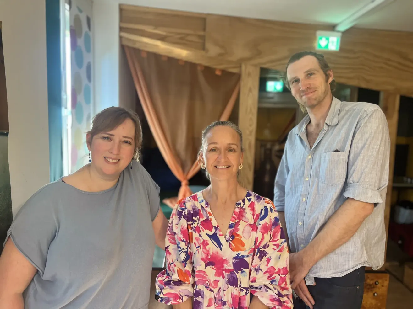 Drei Menschen stehen zusammen und lächeln für ein Foto. Die Frau links trägt ein graues T-Shirt, die Frau in der Mitte eine Bluse mit Blumenmuster und der Mann rechts ein hellblaues Hemd. Hinter ihnen befindet sich eine Holzkonstruktion mit Vorhängen.