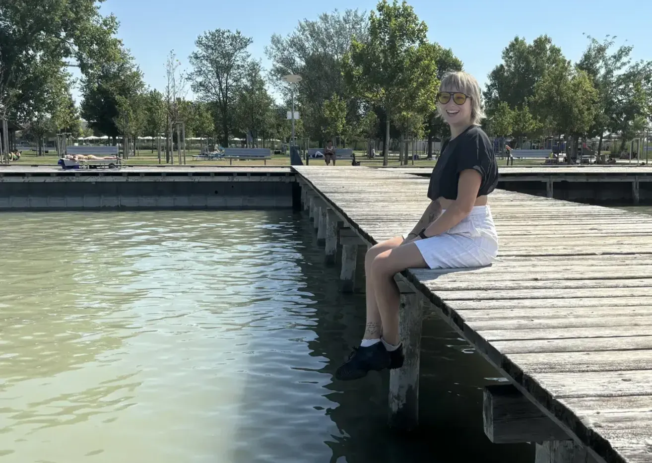 Eine Frau mit Sonnenbrille und schwarzem Shirt sitzt am Rande eines hölzernen Stegs über einem Gewässer und lächelt.