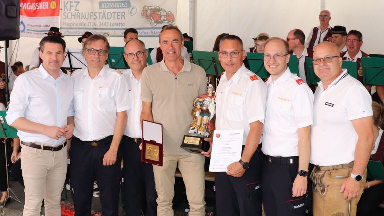Fünf Männer stehen zusammen und lächeln für ein Foto. Der Mann in der Mitte hält eine Trophäe und ein Zertifikat. Die Männer auf beiden Seiten von ihm tragen Uniformen. Hinter ihnen zeigt ein Banner eine Telefonnummer und ein Logo.