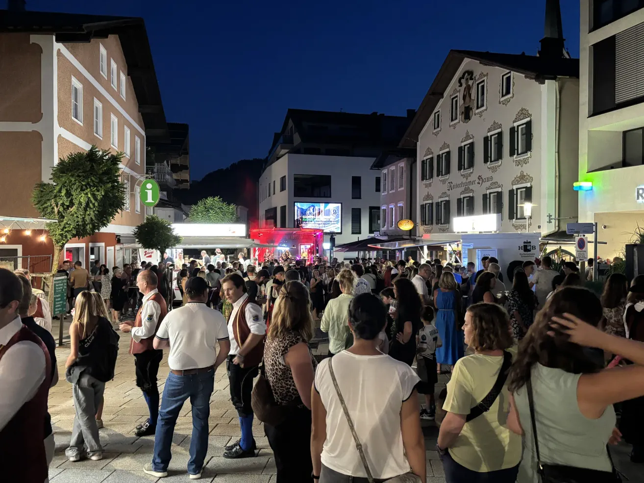 Eine lebhafte Straßenszene bei Nacht mit einer Menschenmenge, die sich um eine Bühne mit roten Lichtern und einer großen Leinwand im Hintergrund versammelt.