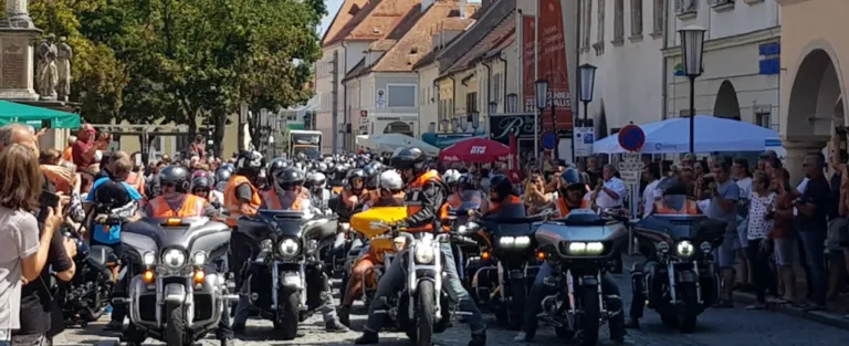 Eine große Gruppe von Motorradfahrern mit Helmen und orangefarbenen Westen versammelt sich auf einer Stadtstraße. Die Motorräder sind aufgereiht und einige sind geparkt. Im Hintergrund befinden sich Gebäude mit Markisen und Straßenlaternen.