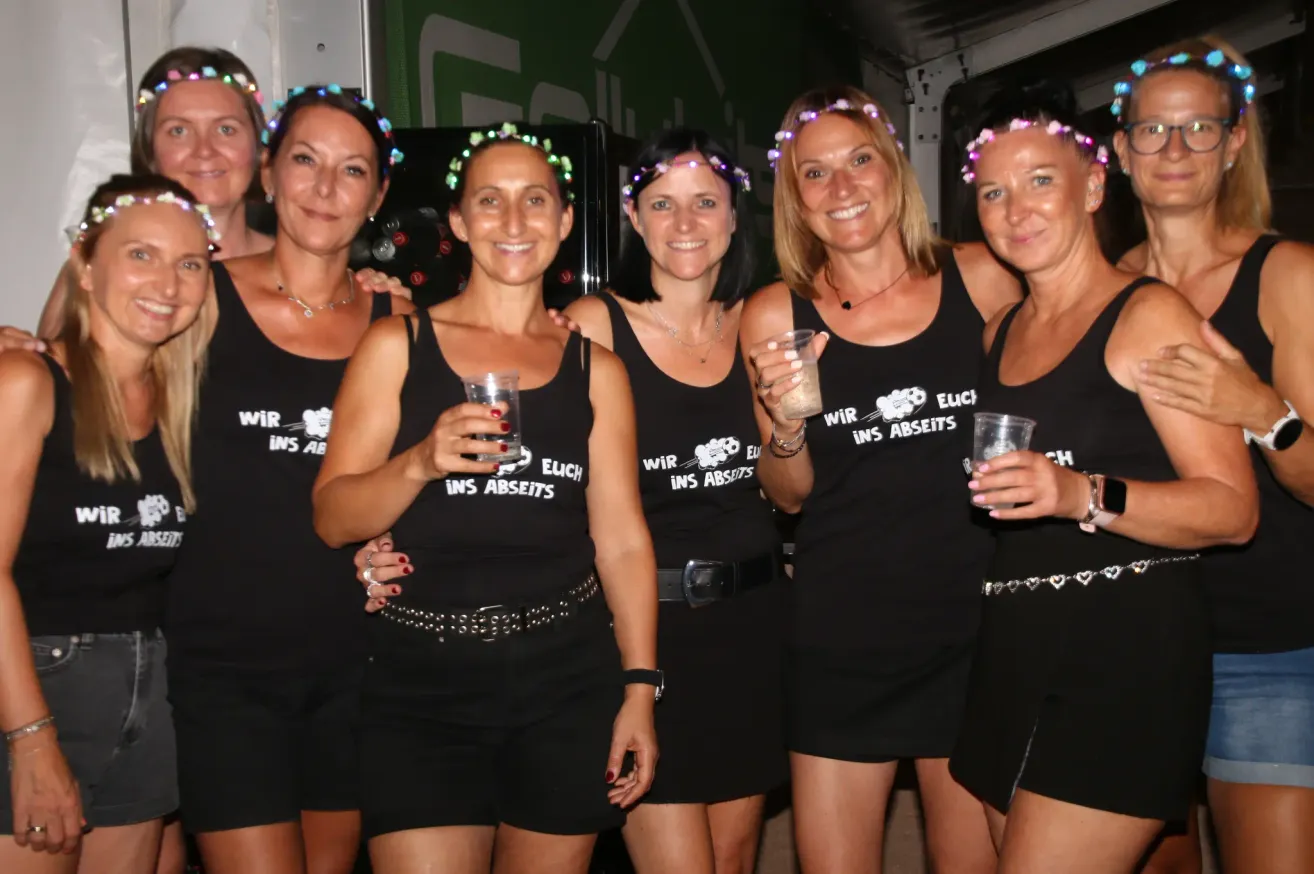 Fünf Frauen in schwarzen Tanktops mit 'Wir ins Abseits' bedruckt posieren für ein Foto, lächeln und halten Getränke. Sie sind mit Blumenkränze geschmückt.