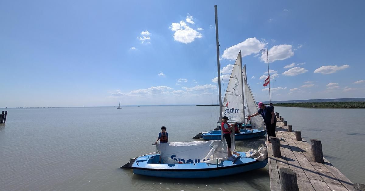 Eine Gruppe von Menschen bereitet sich auf eine Segeltour an einem sonnigen Tag vor. Zwei blaue Segelboote liegen am Pier, auf jedem Boot steht eine Person. Eine weitere Person steht am Pier und hält das Segel des Bootes. In der Ferne segelt ein weiteres Boot auf dem Wasser.