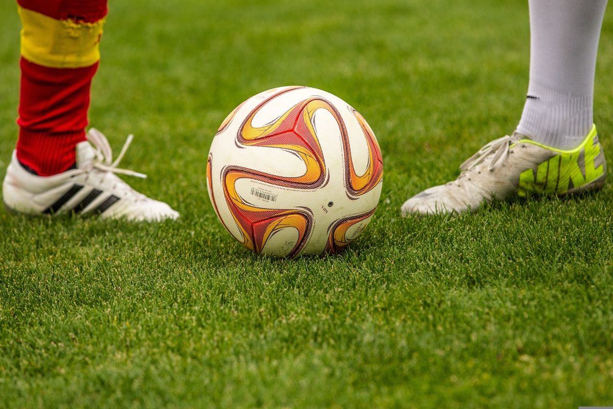 Ein Fußball mit einem markanten Design liegt auf einem üppigen grünen Feld, wobei zwei weiße Schuhe teilweise auf den Seiten sichtbar sind.