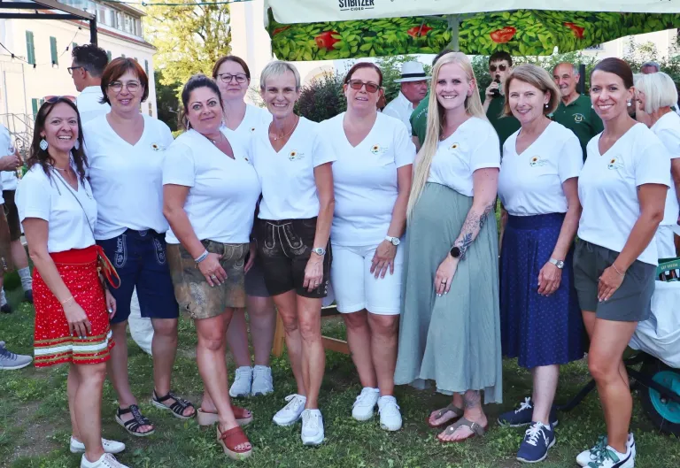 Eine Gruppe von Frauen in passenden weißen Shirts und verschiedenen Hosen, die zusammen auf einer Wiese stehen und für ein Foto lächeln. Hinter ihnen befindet sich ein Baldachin mit grünen Blättern.