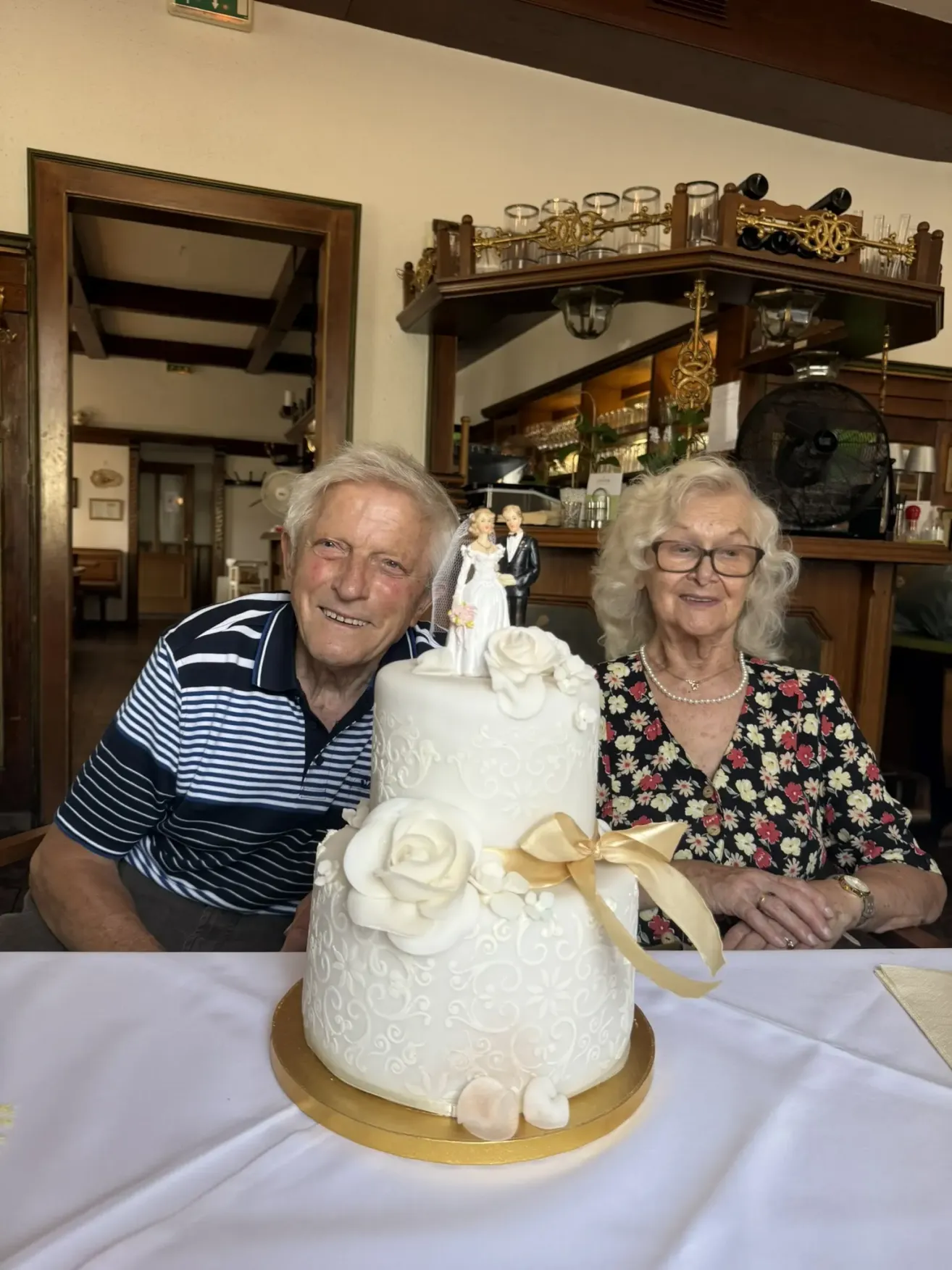 Ein älteres Paar lächelt und posiert mit einem Hochzeitskuchen vor sich. Der Kuchen hat weiße Rosen und eine goldene Schleife. Dahinter befindet sich ein Holzregal mit verschiedenen Gegenständen.