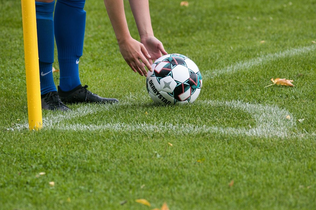 A soccer player in blue shorts and black cleats is holding a Derby Star soccer ball on a grassy field.