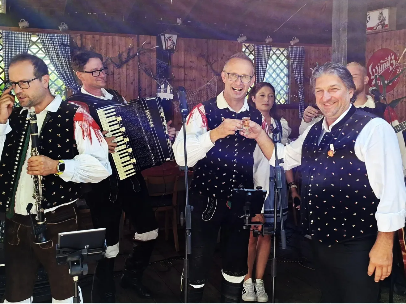 Eine Gruppe von Männern in traditioneller Kleidung tritt auf der Bühne auf, einer spielt Akkordeon und ein anderer hält ein Glas. Eine Frau steht hinter ihnen.