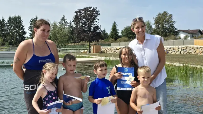 Eine Gruppe von Kindern und zwei Erwachsenen steht am Pool, hält Urkunden und lächelt für ein Foto. Die Frau auf der rechten Seite trägt eine Sonnenbrille auf dem Kopf.