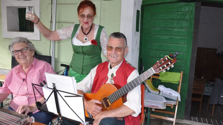 Ein älterer Mann spielt Gitarre und liest Noten, während eine lächelnde Frau in traditioneller Kleidung hinter ihm steht.