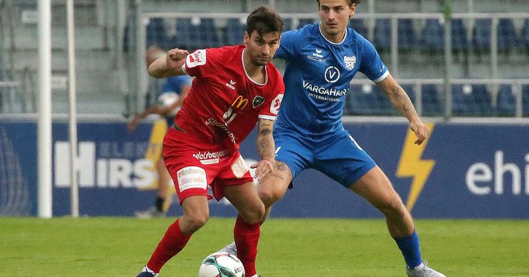 Zwei Fußballspieler in Aktion auf einem Feld, einer in Rot und der andere in Blau, wobei der rote Spieler den Ball schießen will, während der blaue Spieler verteidigt.
