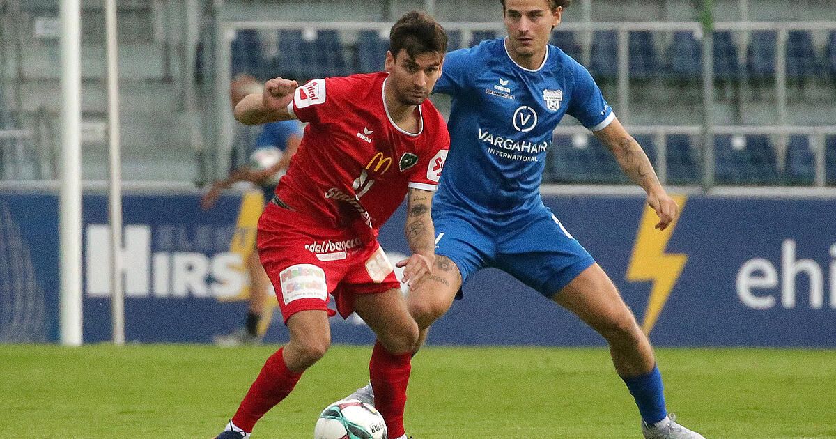 Zwei Fußballspieler in Aktion auf einem Feld, einer in Rot und der andere in Blau, wobei der rote Spieler den Ball schießen will, während der blaue Spieler verteidigt.