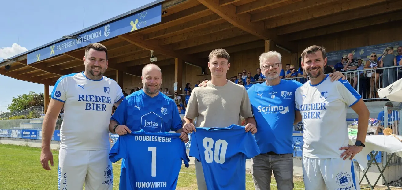 Three men in blue jerseys stand in a stadium, with one holding a jersey with the number 1 and another with the number 80. Behind them, a wooden structure and a crowd of people are visible.