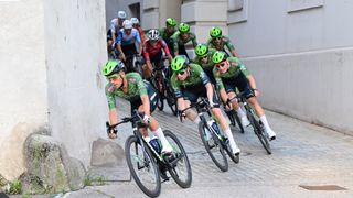 Eine Gruppe von Radfahrern in grünen Helmen und Radsporttrikots fährt auf einer Kopfsteinpflasterstraße. Sie sind in einer Reihe, wobei die Person vorne die Gruppe anführt. Sie tragen alle Sonnenbrillen und Radschuhe.