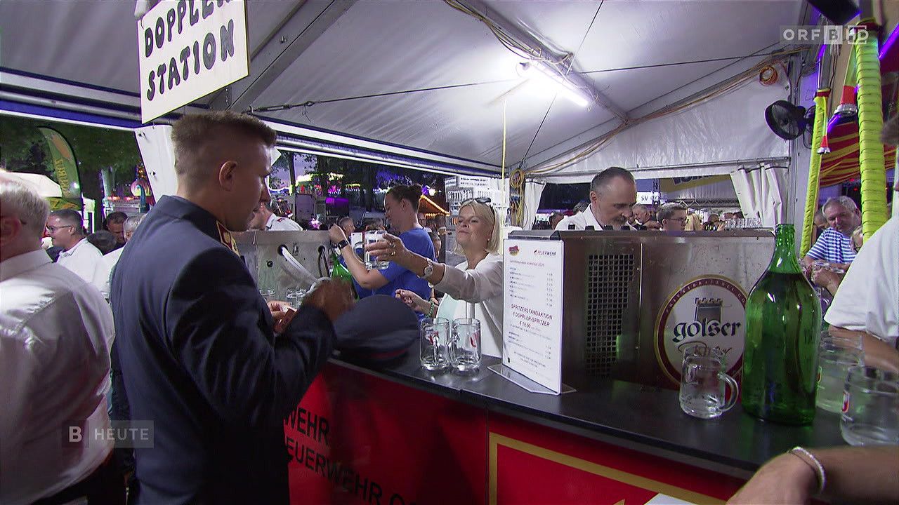 Ein Mann in Uniform serviert einer Frau Getränke an einer Bartheke, mit einer Speisekarte und einem Bierglas davor.