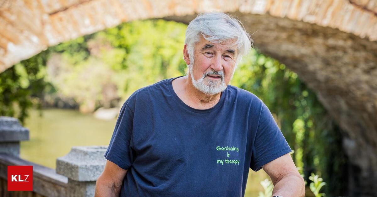 Ein älterer Mann mit weißem Haar und Bart steht unter einem Bogen, trägt ein blaues T-Shirt mit der Aufschrift 'Gartenarbeit ist meine Therapie'.