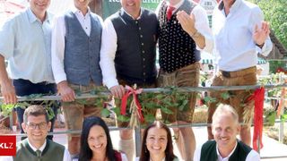 Eine Gruppe von Menschen in traditioneller bayerischer Kleidung, die lächelnd und posierend für ein Foto mit einem festlichen Kranz im Vordergrund.