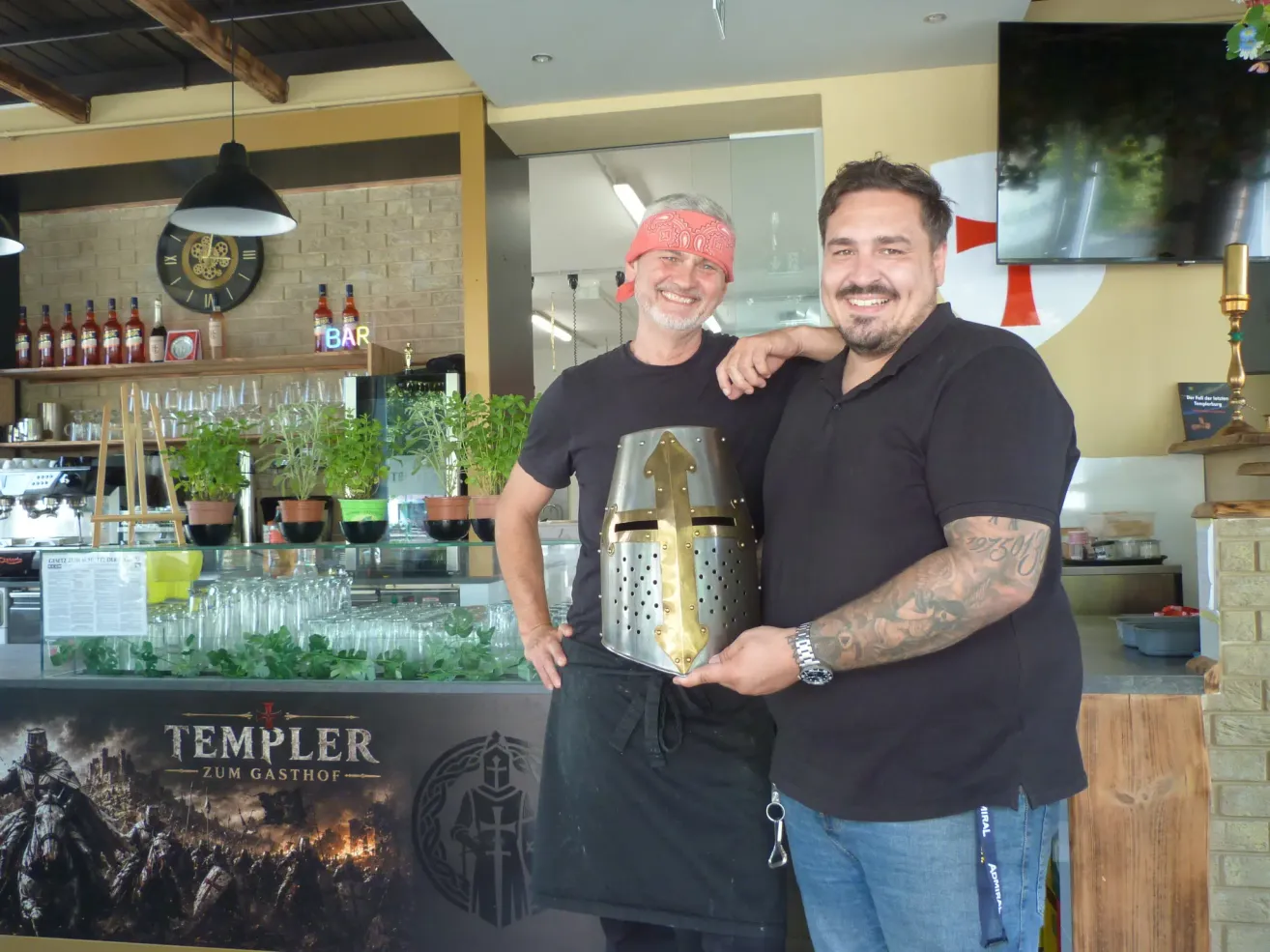 Zwei Männer stehen in einer Bar, einer hält einen Ritterhelm und lächelt für ein Foto. Hinter ihnen steht eine Bartheke mit einem Tempelritter-Banner und Topfpflanzen.