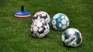 Fünf Fußballbälle mit unterschiedlichen Designs liegen auf einem Rasenfeld, in der Nähe befindet sich ein blauer und roter Kegel.