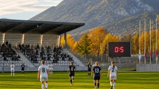 Ein Fußballspiel ist im Gange mit Spielerinnen mit den Nummern 8, 6, 13 und 2 auf dem Feld. Das Spiel wird vor einem Publikum in einem Stadion ausgetragen, wobei die Anzeigetafel 0:5 anzeigt. Im Hintergrund befindet sich ein Berg mit Herbstbäumen.