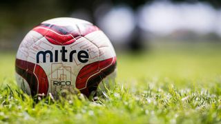 Nahaufnahme eines Fußballs mit den Aufschriften 'Mitre' und 'RICO', der auf einem Rasen liegt.
