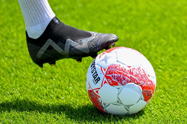 Der Fuß eines Fußballspielers in einem schwarzen und weißen Schuh ist dabei, einen rot-weißen Fußball auf einem grünen Feld zu treten.