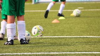 Zwei Fußballspieler in grünen Trikots stehen auf einem Feld mit Fußballbällen und Hütchen. Ein Spieler ist leicht verschwommen.