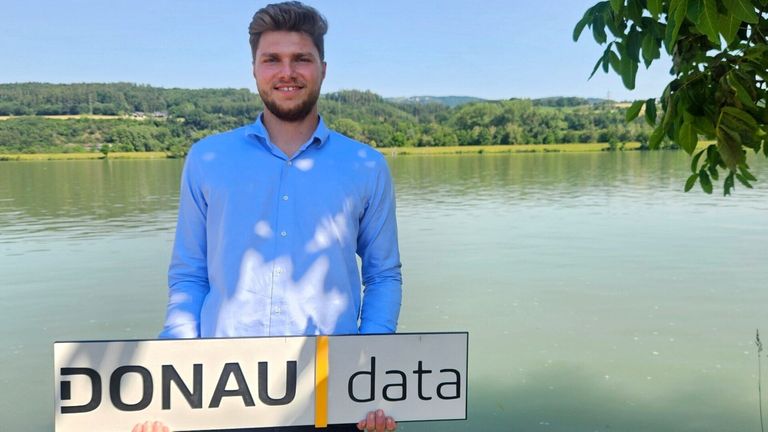 Ein Mann in einem blauen Hemd steht am See und hält ein Schild mit der Aufschrift 'NAU data'. Dahinter ist eine friedliche Landschaft mit Bäumen und Bergen zu sehen.
