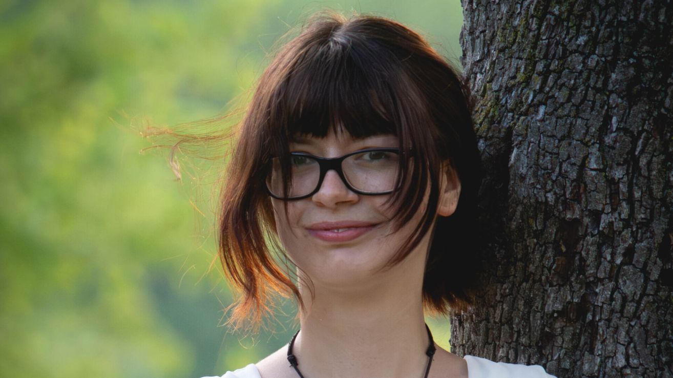 Eine Frau mit kurzen braunen Haaren und Ponyfrisur lächelt und posiert für ein Foto. Sie trägt eine Brille und eine Halskette.