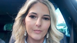 Eine Frau mit blonden Haaren lächelt und trägt Perlenohrringe. Sie hat ein blaues Hemd an.