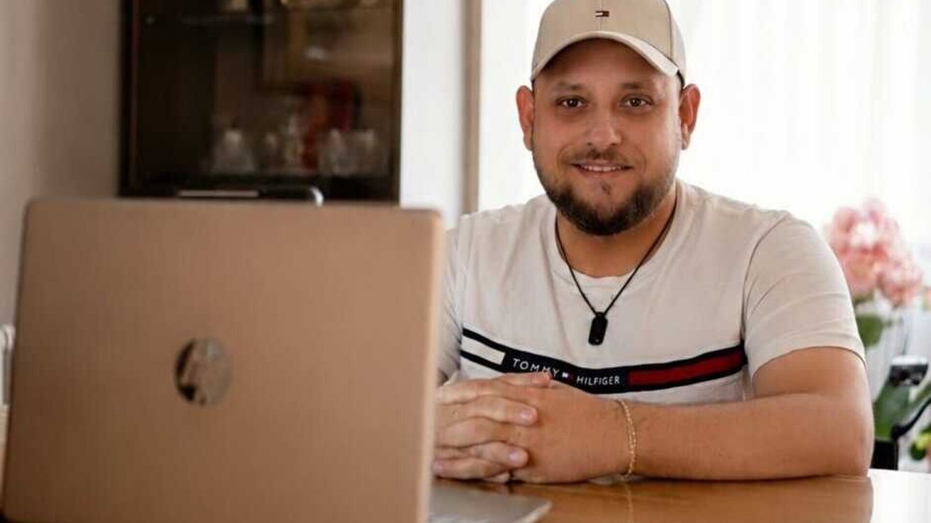 Ein Mann in einem Tommy Hilfiger-Shirt und einer Kappe sitzt mit einem Laptop am Schreibtisch und lächelt.