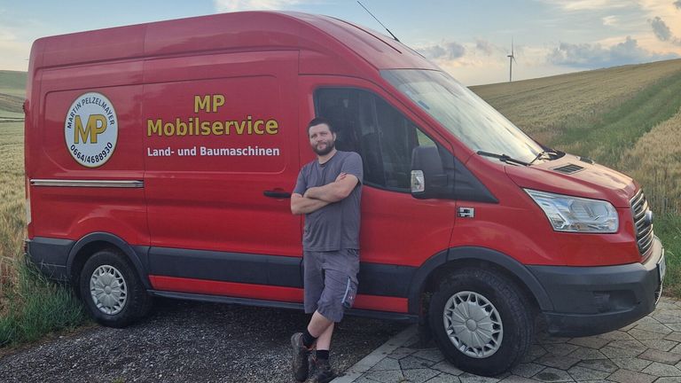 Ein Mann steht neben einem roten Van mit der Aufschrift MP Mobilservice Land- und Baumaschinen an der Seite. Er trägt ein graues T-Shirt und Shorts. Der Van ist auf Kies geparkt.