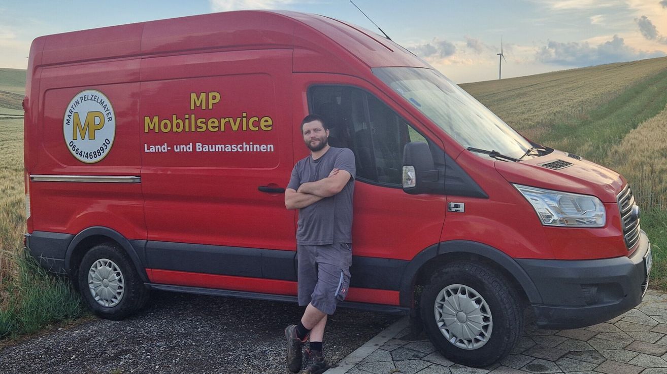 Ein Mann steht neben einem roten Van mit der Aufschrift MP Mobilservice Land- und Baumaschinen an der Seite. Er trägt ein graues T-Shirt und Shorts. Der Van ist auf Kies geparkt.