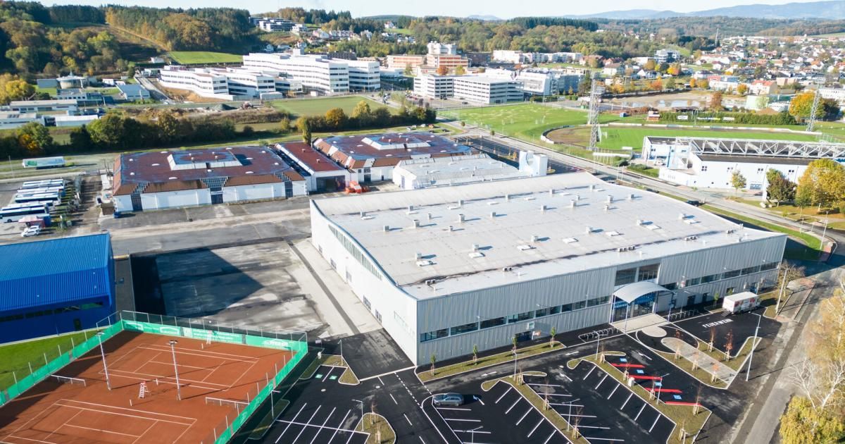 Eine Luftaufnahme eines modernen Industriekomplexes mit mehreren Gebäuden, einem Tennisplatz und einem Parkplatz. Im Hintergrund sind Bäume und Berge zu sehen.