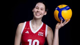 Eine lächelnde Volleyballspielerin in einem roten Trikot mit der Nummer 10 hält einen gelben und blauen Volleyball. Sie trägt eine Halskette und Ohrringe. Das Trikot hat die Wörter 'Burgenland' und 'AUT' darauf. Der Hintergrund ist dunkel.