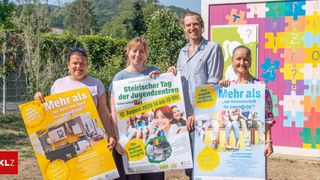 Vier Personen stehen vor einem bunten Gebäude und halten Plakate hoch. Die Plakate haben Text auf Deutsch und Bilder von Menschen. Der Mann in der Mitte hat ein lächelndes Gesicht. Die Frau auf der linken Seite hat eine Sonnenbrille auf dem Kopf. Die Frau auf der rechten Seite hält ein Plakat mit dem Text 'Mehr als nur Gemeinschaft für Jugendliche?'.