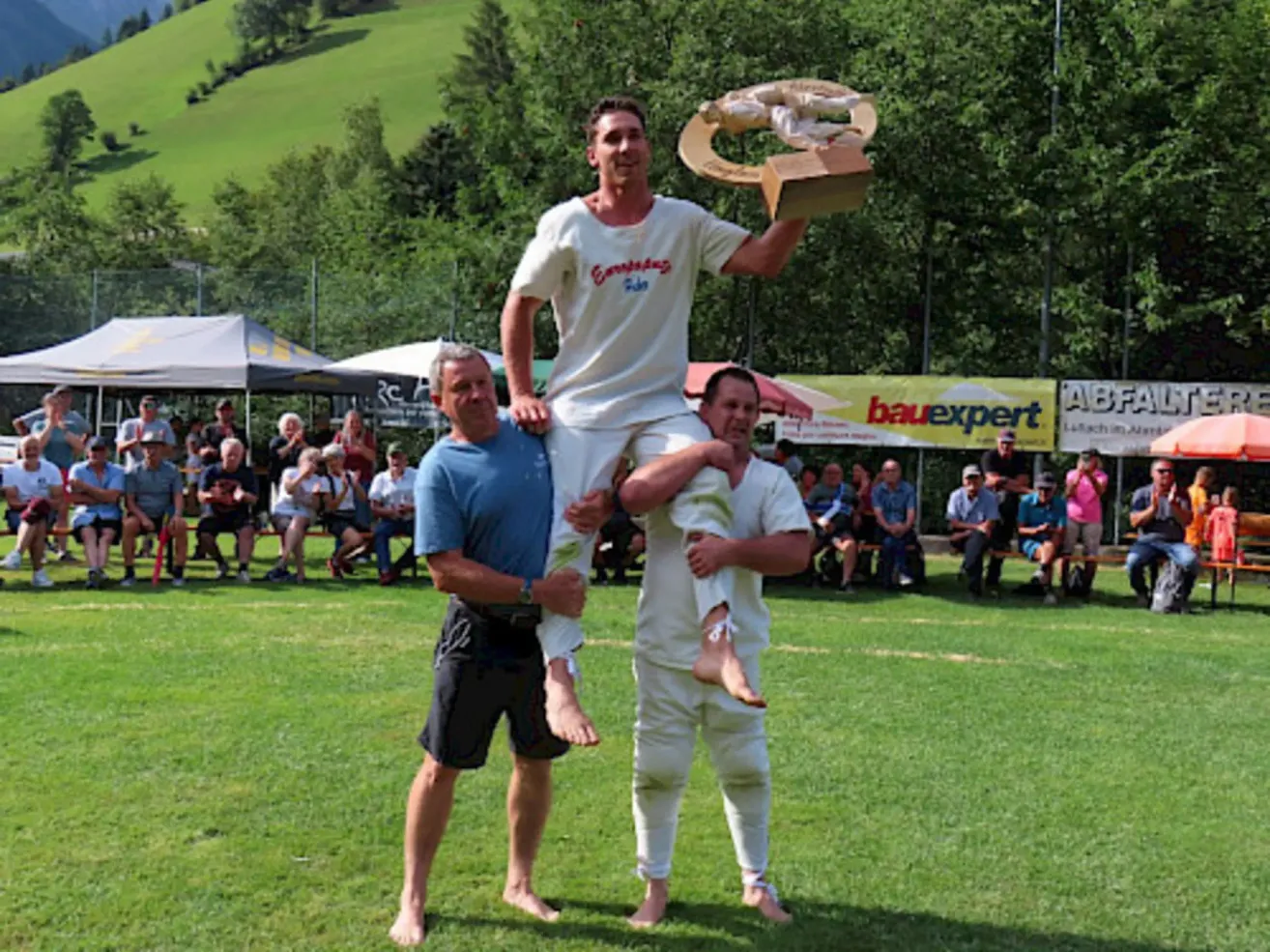 Ein Mann steht auf den Schultern von zwei anderen und hält eine Trophäe in einem Grasfeld, mit Zuschauern im Hintergrund.