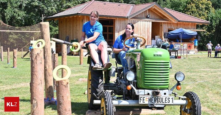 Zwei Frauen sitzen auf einem grünen Traktor, eine fährt und die andere sitzt auf dem vorderen Sitz. Sie befinden sich in einem Grasbereich mit Holzpfosten und einem Holzhaus im Hintergrund.