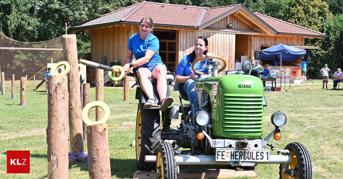 Zwei Frauen sitzen auf einem grünen Traktor, eine fährt und die andere sitzt auf dem vorderen Sitz. Sie befinden sich in einem Grasbereich mit Holzpfosten und einem Holzhaus im Hintergrund.