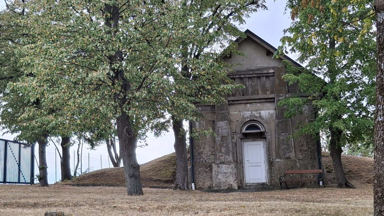 Ein altes Steingebäude mit einer weißen Tür steht auf einem Feld, flankiert von zwei Bäumen.