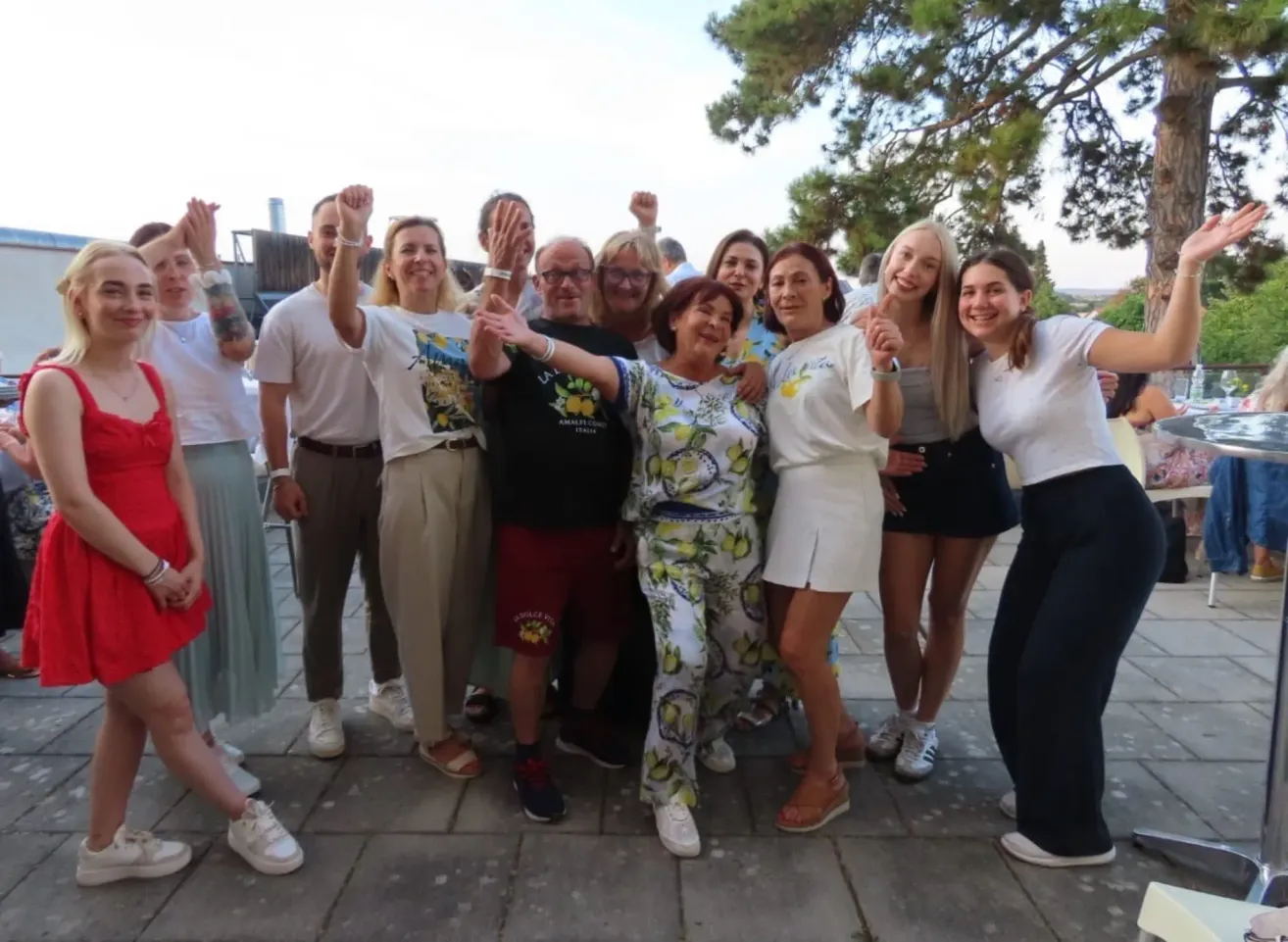 Eine Gruppe von Menschen, darunter Erwachsene und Kinder, posiert für ein Foto im Freien. Sie stehen eng beieinander, lächeln und einige heben ihre Hände. Im Hintergrund befinden sich Bäume und ein klarer Himmel.