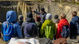 Eine Gruppe von Menschen, einschließlich Kindern, versammelt sich um eine Karte und Steine, möglicherweise auf einer Geologie-Tour. Der Mann in der Mitte zeigt auf etwas auf der Karte.