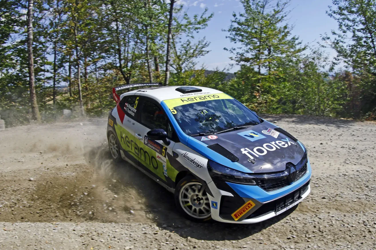 A rally car with the number 5 on the hood is driving on a dirt road with trees in the background.